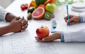 Hand holding apple and other hand writing on clipboard with patients hand folded on other side of table.